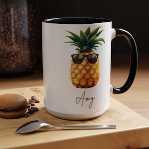 Op de afbeelding: Witte keramische mok met een zwarte rand en handvat. De mok heeft een kleurrijke illustratie van een ananas met een zonnebril en de naam "Amy" in zwarte cursieve letters.