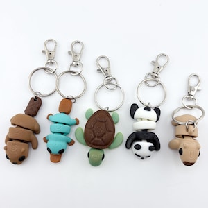 Op de afbeelding: Vijf handgemaakte sleutelhangers van polymeerklei met dierontwerpen. De sleutelhangers omvatten een beer, een schildpad, een panda en twee andere dierfiguren. Elke sleutelhanger heeft een zilverkleurige sluiting en ring.