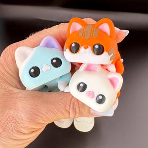 May include: Three colorful cat figurines. One is light blue with a purple head, another is orange with brown stripes, and the third is white with pink ears. Each has large black eyes and a cute expression. The figurines are stacked together.