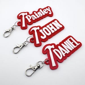 Op de afbeelding: Drie rode sleutelhangers met witte letters die "Paisley", "John" en "Daniel" spellen. Elke sleutelhanger heeft een zilveren sluiting. De sleutelhangers zijn een gepersonaliseerd accessoire.