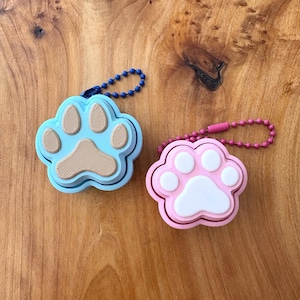 Può includere: Due portachiavi a forma di zampa. Uno è azzurro con cuscinetti marroni e una catena di perline blu scuro. L'altro è rosa con cuscinetti bianchi e una catena di perline rosa. Entrambi sono su una superficie di legno.