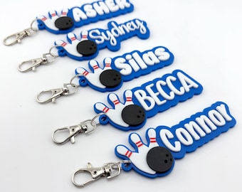 May include: Five silicone keychains featuring a bowling theme with a white bowling pin and a black bowling ball. Each keychain has a silver metal lobster clasp and displays a unique name in white raised lettering: Asher, Sydney, Silas, Becca, and Connor.