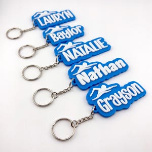 May include: A collection of blue keyrings, each with a name in white lettering and a swimmer graphic. The names, such as Lauryn and Nathan, are attached to silver keyrings. Ideal for swimmers.