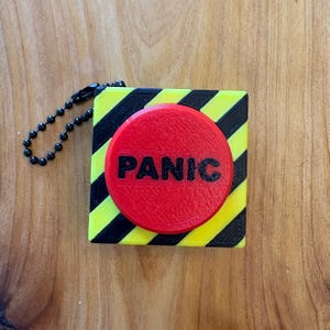 Może przedstawiać: Czarno-żółte pudełko z czerwonym przyciskiem, na którym widnieje napis "PANIC" czarnymi literami. Pudełko ma czarny łańcuch.