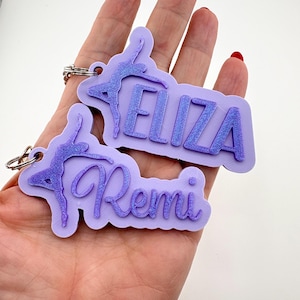 May include: Two lavender keychains with a gymnast silhouette and names "Eliza" and "Remi" in cursive. The gymnast and names have a glittery, darker purple accent. Each keychain has a silver keyring attached.