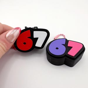 May include: Two keychains with the numbers 67. One keychain has a red 6 and a white 7, the other has a purple 6 and a pink 7. Both keychains are black with a small chain.