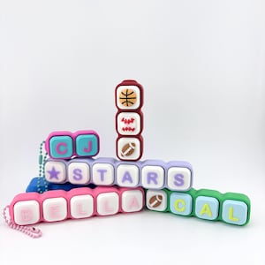 May include: A collection of silicone bead keychains featuring colorful rectangular blocks with letters and sports icons. The blocks display the names CJ, STARS, BELLA, and CAL, alongside graphics of a basketball, baseball, and football, all attached to a beaded chain.