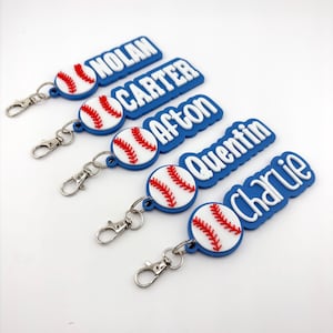 May include: Baseball-themed keychains with names: Nolan, Carter, Afton, Quentin, and Charlie. Each keychain features a baseball design with red stitching and a blue background. The names are in white, raised lettering.