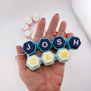 以下が含まれることがあります： JOSHとCAMの名前が入った六角形のビーズの連結チェーン2本。上のチェーンは濃い青のビーズに白い文字、下のチェーンは水色のビーズに黄色い文字が施されています。どちらのチェーンにも白いボールチェーンが付いています。