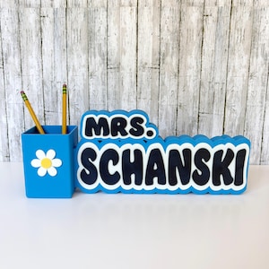 May include: A blue desk tidy set with a daisy accent and the name "MRS. SCHANSKI" in black and white. The set includes a pencil holder with two pencils.