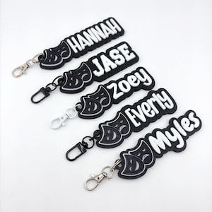 May include: Black and white keychains with names Hannah, Jase, Zoey, Everly, and Myles. Each keychain features a theater mask design and a silver-colored clasp.