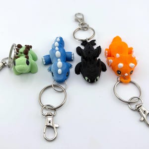 May include: Five colorful dragon-shaped keychains. The keychains are green, blue, black, and orange. Each dragon has a silver keyring and a lobster clasp. The dragons are lying down with their wings spread.