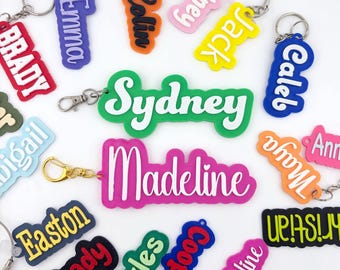 Op de afbeelding: Een verzameling kleurrijke, op maat gemaakte naamsleutelhangers met diverse namen zoals Sydney, Madeline, Brady, Emma, Colin, Jack, Caleb, Abigail, Maya, Ann, Easton, Myles, Cooper en Kristian, elk voorzien van een metalen ring of karabijnhaak.