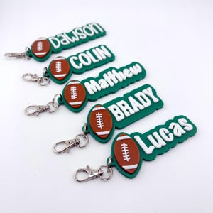 May include: Green and white personalized football keychains with names like Dawson, Colin, Matthew, Brady, and Lucas. Each keychain features a brown football with white stitching and a silver clasp.