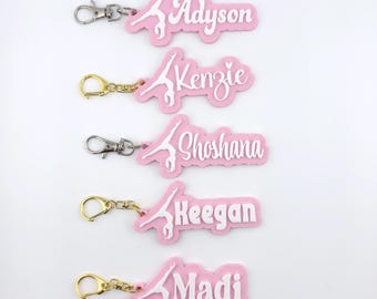 Op de afbeelding: Vijf roze acryl sleutelhangers met een witte silhouet van een turner in handstand. Elke sleutelhanger toont een unieke naam in witte schrijf- of blokletters: Adyson, Kenzie, Shoshana, Keegan en Madi, bevestigd aan metalen sluitingen.
