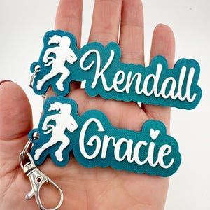May include: Two teal keychains with white text that read "Kendall" and "Gracie." Each keychain features a white silhouette of a person running. The keychains have silver-colored clasps.
