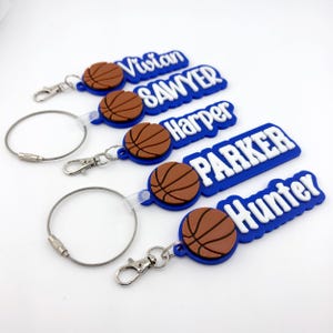 Puede incluir: Cinco llaveros de silicona con un emblema de baloncesto marrón y letras blancas en relieve. Los nombres Vivian, Sawyer, Harper, Parker y Hunter aparecen sobre fondos azules. Cada llavero incluye un cierre de mosquetón o un aro metálico.