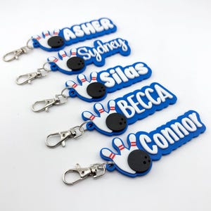 Puede incluir: Una colección de llaveros personalizados con temática de bolos. Cada llavero presenta un fondo azul con un nombre blanco, un diseño de bolos y una bola de bolos negra. Los llaveros tienen cierres plateados.