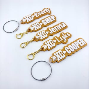 May include: Five rectangular mustard yellow keychains with white raised lettering and a white arrow graphic. Each keychain features the text Everly, Shep, Jenna, Lyla, or Cooper, paired with the letters XC. Each includes a metal ring attachment.