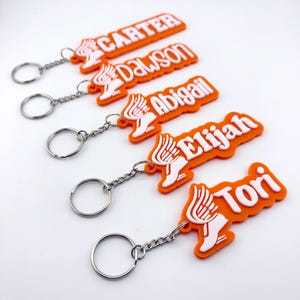 May include: Five orange and white silicone keychains, each featuring a winged shoe icon and a unique name in white lettering: Carter, Dawson, Abigail, Elijah, and Tori. Each keychain includes a silver metal split ring attached by a chain.
