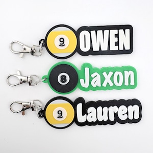 May include: Three personalized keychains with silver clasps. Each keychain features a pool ball design with the numbers 8 or 9, and a name: Owen, Jaxon, and Lauren. The names are in white, with the pool ball designs in black, yellow, and green.