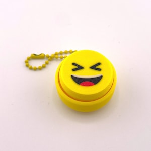 May include: A yellow keychain featuring a smiling emoji face. The emoji has black eyes and a wide, red mouth.