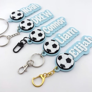 May include: A collection of personalized soccer-themed keychains. Each keychain features a soccer ball design and a name in white lettering on a light blue background. The keychains have various metal hardware.