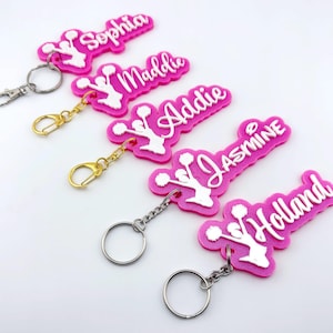 May include: Pink personalised keychains with white text spelling out names: Sophia, Maddie, Addie, Jasmine, and Holland. Each keychain features a cheerleader design and a metal clasp or keyring.