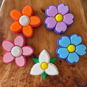 May include: A collection of five colorful flower-shaped objects. The flowers are in shades of orange, pink, purple, and blue, each with a white or yellow center. The flowers are arranged on a wooden surface.