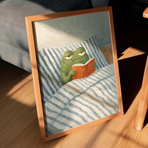 May include: A framed print depicting a green frog in bed, reading a book with a grumpy expression. The bedding has blue and white stripes. The frame is light brown. The print is leaning against a grey sofa and wooden chair.