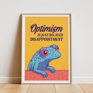 Op de afbeelding: Een gele poster met een blauwe kikker met roze ogen en de tekst "Optimisme is gewoon uitgesteld teleurstelling."