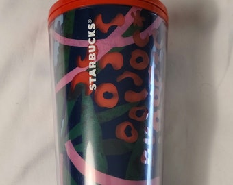 May include: A clear plastic tumbler with a red lid and a purple straw. The tumbler has a blue background with a pink and green floral pattern. The text "STARBUCKS" is printed on the side of the tumbler.