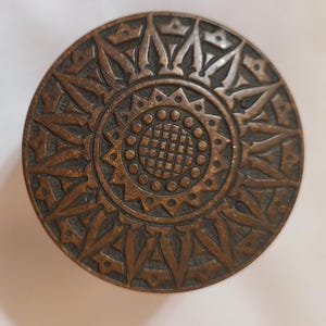 Puede incluir: Un objeto decorativo redondo de color bronce con un intrincado diseño de sol. La parte central presenta un patrón circular con pequeños puntos, rodeado por un anillo de pétalos estilizados y formas puntiagudas que irradian hacia afuera.