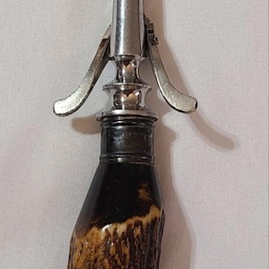 May include: A vintage carving fork with a stainless steel head and a handle made of textured brown antler. The fork has two tines and a lever mechanism. A classic kitchen utensil.