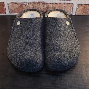 May include: A pair of dark gray felted wool clogs with a rounded toe and an open back. Each clog features a small, circular metallic rivet on the outer side near the opening. The interior is lined with a light-colored, textured material.