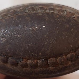 May include: Close-up of a dark brown, oval-shaped metal object. The object has a textured surface and a decorative border of small, rounded bumps. The metal appears aged and possibly antique.