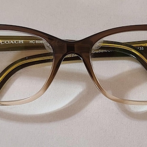 Puede incluir: Gafas Coach marrones con montura degradada, que va del beige claro al marrón oscuro. Las patillas son marrones lisas con detalles dorados. El número de modelo HC 8085 es visible.