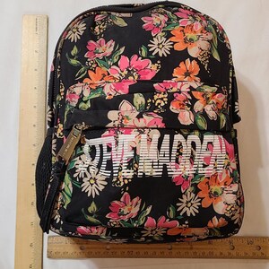 May include: Black floral backpack with white "Steve Madden" text. The backpack has a zipper closure and a mesh pocket on the side.