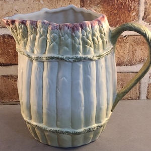 May include: A ceramic pitcher with a light blue body and a decorative asparagus design around the top. The handle is green, and the rim has a pink and green trim. The pitcher has vertical ridges and a textured appearance.