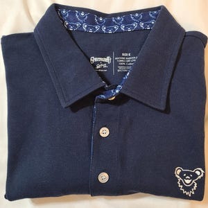 Op de afbeelding: Marineblauwe poloshirt met een wit geborduurd berenlogo links onder. De kraag en de binnenste knooplijst hebben een patroon. De shirt heeft twee witte knopen en een label met de tekst "Grateful Dead".