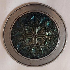 Könnte beinhalten: Ein runder, silberfarbener Untersetzer mit einem erhabenen, grün-blauen Blumendesign in der Mitte.