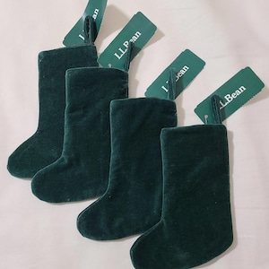 May include: Four dark green velvet Christmas stockings with attached green tags that read "L.L.Bean". The stockings are arranged in a row, with the tags angled upwards. The stockings are a solid color and have a soft, plush appearance.