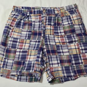 May include: A pair of shorts featuring a multi-coloured patchwork plaid pattern. The design consists of various squares of fabric in shades of blue, white, red, yellow, and green. The shorts include a waistband with belt loops and a central button.