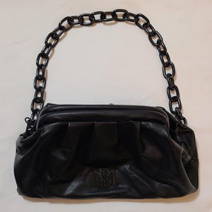 May include: Black leather handbag with a chain handle. The bag has a gathered design and a logo on the front. The chain handle is made of large, dark links. The bag is a clutch style.