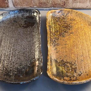 Puede incluir: Dos fuentes de cerámica con formas orgánicas. Una fuente es de color gris oscuro con una textura moteada, y la otra es de color marrón dorado con una textura similar. Las fuentes tienen un aspecto rústico y artesanal.