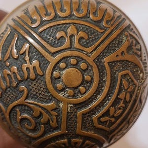 May include: Close-up of a bronze-colored, ornate button with a textured surface. The button features a central circular design with a series of smaller circles, surrounded by decorative patterns and the text "17 mm".