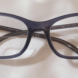 May include: A pair of gray eyeglasses with a rectangular frame. The frame has a subtle cat-eye shape. The glasses have clear lenses and a metal nose bridge.