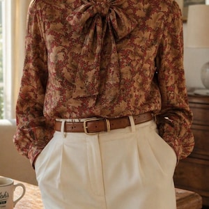May include: A long-sleeved blouse with a floral pattern in shades of brown and red, featuring a bow tie neckline. The blouse is paired with cream-colored pleated trousers and a brown leather belt with a gold buckle. The outfit is completed with a coffee mug.
