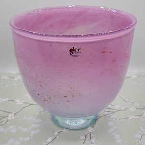 Puede incluir: Un cuenco grande y redondo de cristal rosa con una amplia abertura. El cuenco tiene un diseño moteado con motas doradas y una base azul claro. El cuenco tiene una etiqueta que dice "der", "BERGSEIE" y "HANDMADE IN ICELAND".