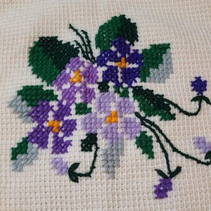 May include: Close-up of a cream-colored, textured fabric with a cross-stitched floral design. The design features purple, violet, and yellow flowers with green leaves and stems. The cross-stitch creates a detailed, textured appearance.